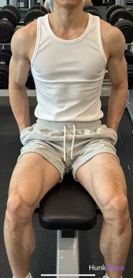 Seated on a black weight bench, torso upright with hands resting on the edges beside hips and legs s
