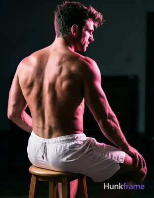 A shirtless adult man with sculpted upper back muscles and defined latissimus dorsi sits on a wooden