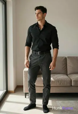 A handsome adult man with short black hair and clean-shaven face stands casually in a modern apartme