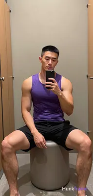 Asian man with short black buzz cut and light stubble sits on a cylindrical ottoman in a gym locker 