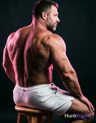A big hairy stocky adult man with short dark hair and clean-shaven jaw sits shirtless on a wooden st