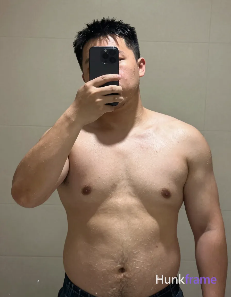 A big hairy stocky masculine man stands shirtless in an indoor bathroom or shower area with beige tiled walls, torso angled slightly right; He holds a black smartphone with his left hand up to face, partially obscuring the eye region and lower jawline against cheek/eye area; Broad shoulders display defined pectoral muscles and visible abdominal definition on light to medium tan skin with visible sheen from moisture or sweat; Soft directional lighting creates highlights on wet skin while high contrast separates him from the matte background; Vertical portrait crop focusing on upper torso, centered composition cropped from mid-chest up to forehead where head is partially cut off at top left; single clean uninterrupted photograph with a cohesive editorial composition and plain continuous surrounding surfaces