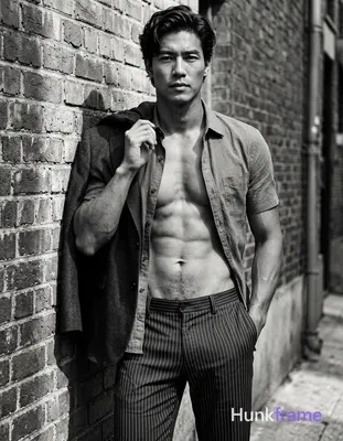 A handsome medium-dark skinned man with dark wavy hair styled back stands leanin