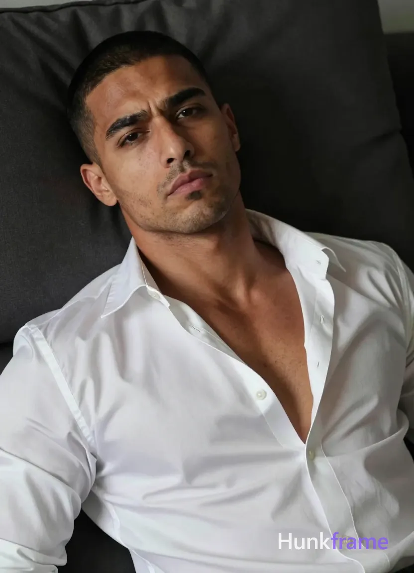an anonymous figure man with light to medium tan skin texture and pores, short black buzz cut, light stubble on chin; Lying supine on back in a dark bedroom setting, head tilted slightly left against pillow shoulders relaxed; Wearing white satin button-down shirt with wide collar and deep V-neck exposing upper chest only; Soft directional lighting from front-left creates subtle shadows on right side of face; High-resolution skin detail, minimalist composition tight framing on expressive grimacing face and torso; Athletic photography style, same aesthetic theme and photo feel as the reference but a different man in a different pose with distinct body proportions, Middle Eastern man, tan skin, dark beard shadow, muscular but natural proportions; single clean uninterrupted photograph with a cohesive editorial composition and plain continuous surrounding surfaces