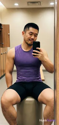 Asian man with short black buzz cut and light stubble sits on a cylindrical ottoman in a gym locker 