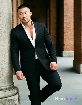 Asian man with short buzz cut and light stubble stands in three-quarter profile leaning against a st