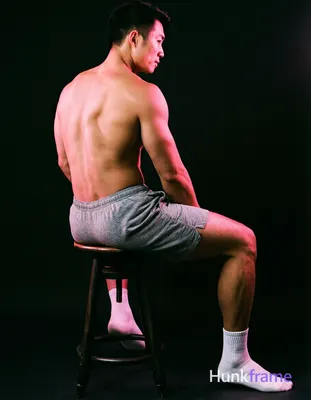 Asian man seated on a dark stool, torso angled away with left leg bent and right extended forward; S