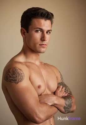 A muscular young man with short brown hair stands shirtless against a neutral ba