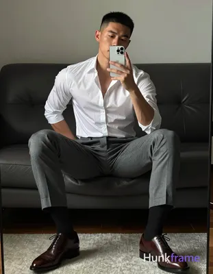 Asian man seated on dark leather sofa, legs spread knees bent taking mirror selfie with white smartp