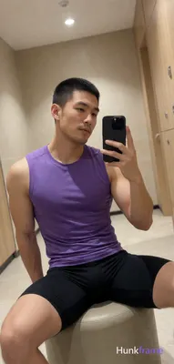 Asian man with short black buzz cut and light stubble sits on a cylindrical ottoman in a gym locker 