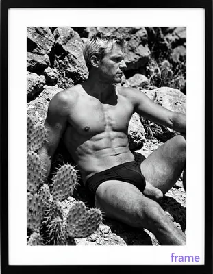 A lean white man with short light-colored hair sits contemplatively on rocky ground, torso turned sl
