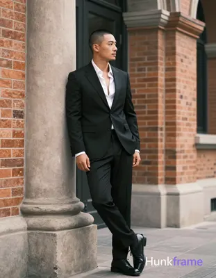 Asian man with short buzz cut and light stubble stands in three-quarter profile leaning against a st