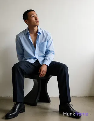 Asian man with short buzz cut dark brown hair and slight upper lip stubble seated on a black sculptu