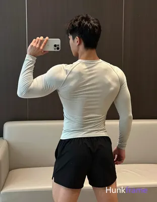 A muscular masculine man stands facing away from the camera toward a window or m