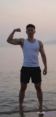 Asian man with medium-dark skin and short dark hair stands in shallow beach water at dusk, flexing r