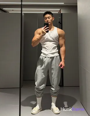 Asian man with short black buzz cut and light stubble stands full-body in a gym mirror, wearing whit