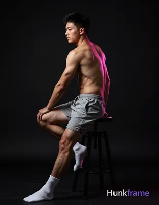 Asian man seated on a dark stool, torso angled away with left leg bent and right extended forward; S