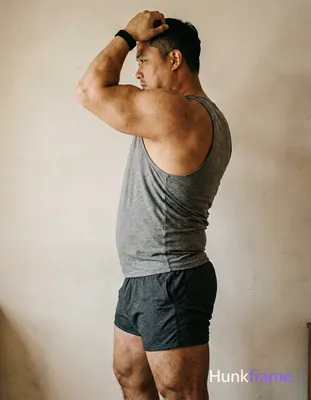 A big hairy stocky masculine man with bear energy, standing in profile with left arm raised behind h