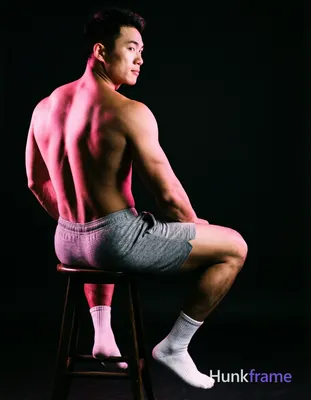 Asian man seated on a dark stool, torso angled away with left leg bent and right extended forward; S