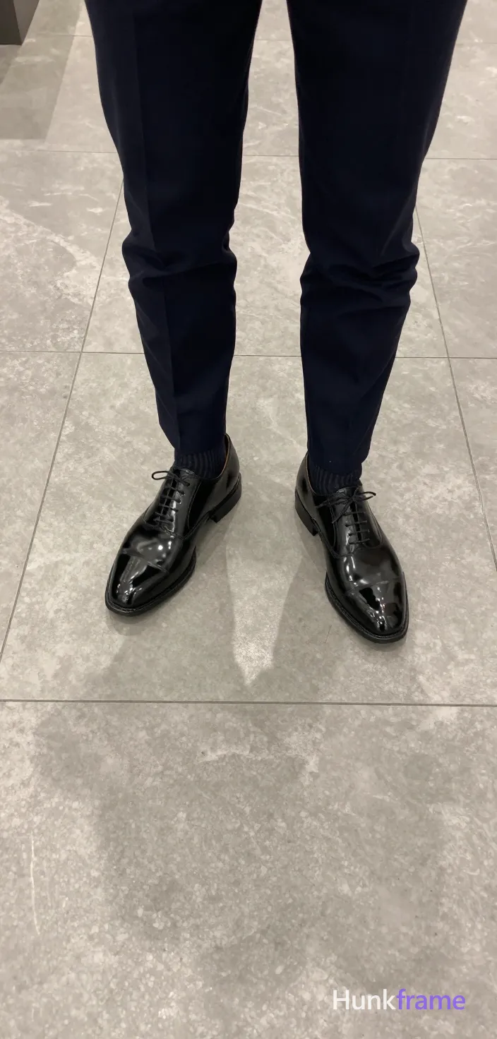 Top-down fashion editorial crop focusing on luxury footwear and tailored trousers. A handsome masculine man stands centered with legs slightly apart, viewed from above showing only lower body cropped at thighs and feet. He wears dark navy trousers paired with black ribbed socks featuring a faint script logo on the right sock, finishing in shiny black patent leather lace-up oxford shoes reflecting soft overhead ambient lighting. The setting is an indoor space with light gray marble or stone tile floor displaying visible grout lines. High-contrast specular reflections highlight the shoe texture against the neutral backdrop., a single uninterrupted photograph only, no written words or typography anywhere in frame, no captions, no app UI, no social-media post layout, no screenshot borders
