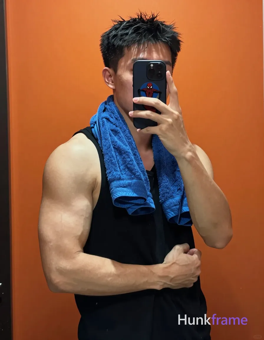 Asian man with light to medium tan skin and short black hair, slightly tousled straight texture, clean-shaven face; Standing facing a mirror in an indoor bathroom or dressing room with orange wall background; He is shirtless adult bare chest showing broad shoulders, defined pectorals, large biceps with prominent veins, toned forearms; Wearing a black sleeveless tank top (no shirt) and blue towel draped around neck; Holding black smartphone with blue Spider-Man case up to face for mirror selfie in right hand; left arm bent at elbow displaying muscles, left hand making loose fist near chest level; High contrast lighting from phone camera flash creating harsh highlights on skin and muscles; Portrait composition centered in mirror reflection, vertical 13:19 aspect ratio, clean negative space; single clean uninterrupted photograph with a cohesive editorial composition and plain continuous surrounding surfaces