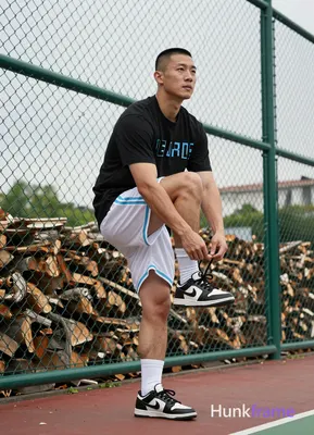 Asian man with short black buzz cut and clean-shaven face, standing casually near a green chain-link