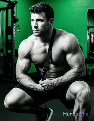 A muscular adult man with dark short slightly wavy hair and clean-shaven face crouches in a gym sett