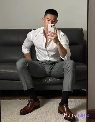 Asian man seated on dark leather sofa, legs spread knees bent taking mirror self