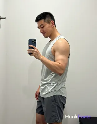 Asian man with short black buzz cut and light stubble smiles while taking a mirror selfie, standing 
