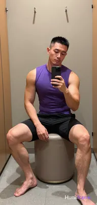 Asian man with short black buzz cut and light stubble sits on a cylindrical ottoman in a gym locker 