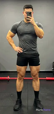 A very muscular adult man stands full-body facing the camera in a gym mirror sel