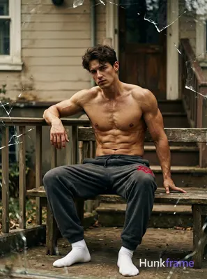A handsome masculine man sitting on a wooden bench with one hand resting on a railing, leaning sligh