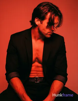 Adult man seated in a moody studio with solid red backdrop, shirtless under an o