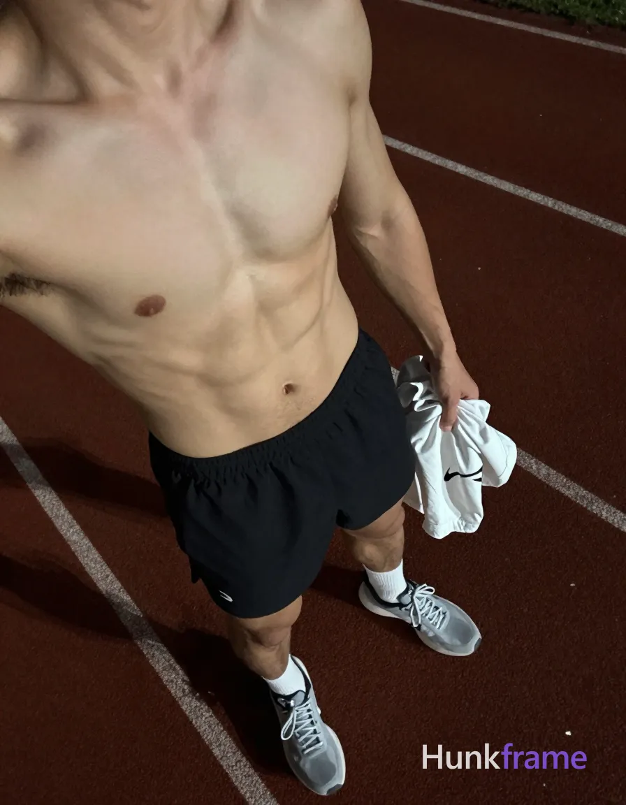 High-angle POV fashion editorial shot of a handsome adult man standing on an outdoor running track at night, torso angled toward camera. He is shirtless with defined abdominal musculature and thick thighs visible from lower ribs to upper abdomen. Left hand holds a crumpled white Nike-branded garment near hip; right arm not visible. Wearing black athletic shorts with small white logo on left thigh, white crew socks, grey and white running shoes. Dark reddish-brown rubberized track surface with faded white lane lines under low-key ambient lighting. Soft directional highlights sculpt skin texture against slightly desaturated tones. Grainy photo aesthetic with shallow depth of field focusing on torso and feet. Intimate post-workout mood occupying right and bottom frame portions; head cropped out. Include exactly one visible wordmark, spelled exactly "HF", as a small garment print; preserve its appropriate typography and scale. No other visible text, logo, caption, username, hashtag, comment, watermark, or UI chrome., a single uninterrupted photograph only, no written words or typography anywhere in frame, no captions, no app UI, no social-media post layout, no screenshot borders