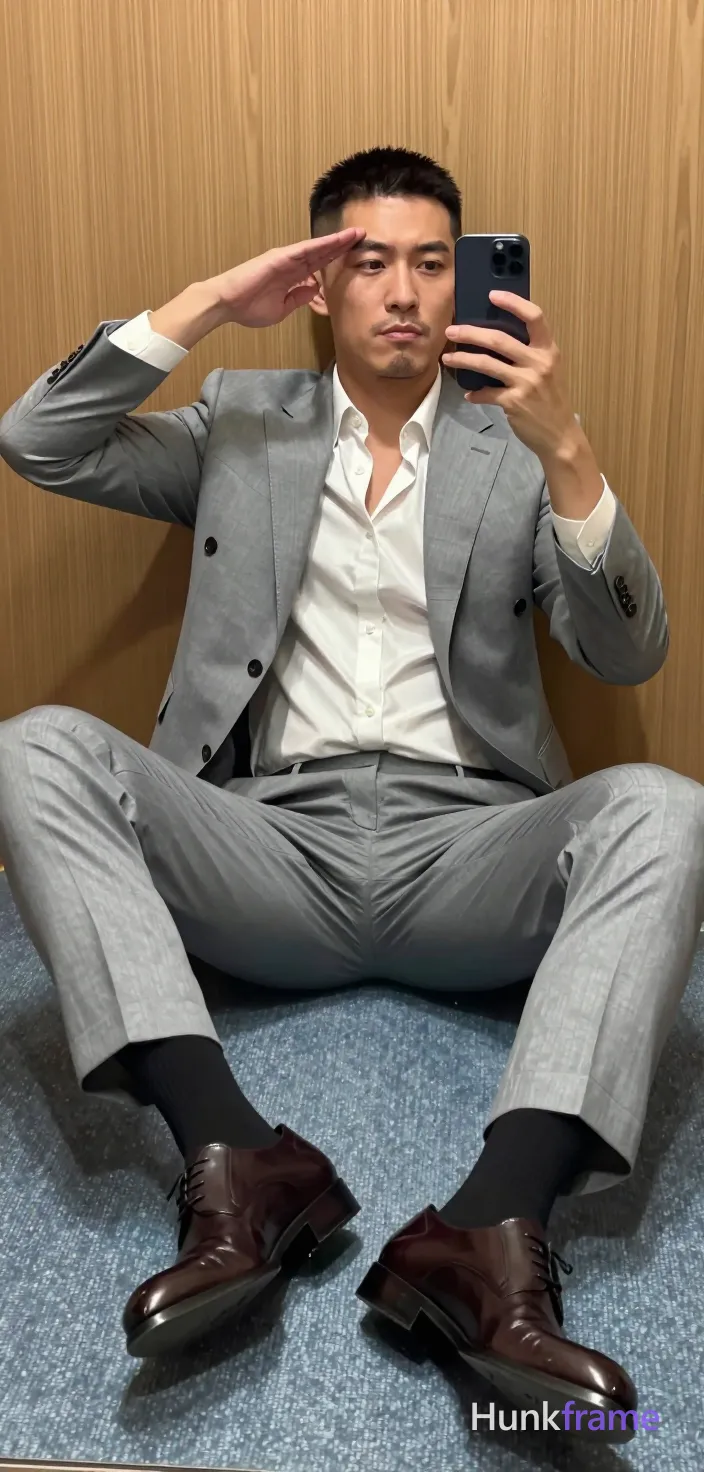 Asian adult man with short black buzz cut and light stubble, neutral expression saluting his forehead with right hand while holding dark smartphone in left for mirror selfie; Seated on light blue carpeted floor legs spread wide X-shape torso leaning back against wood paneling wearing open white dress shirt revealing upper chest cleavage sleeves rolled up paired with light grey single-breasted suit jacket and matching tailored trousers black socks dark brown leather shoes soft indoor lighting vertical portrait composition confident stylish mood polished anime 2.5D next-gen game art style; single clean uninterrupted artwork with a cohesive composition and plain continuous surrounding surfaces