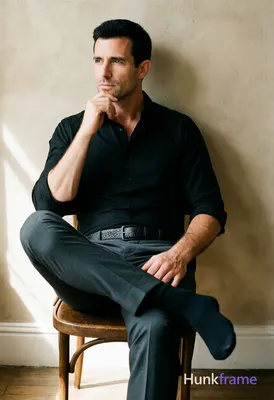 Adult man with short black hair and clean-shaven face sits in a wooden chair, leaning back against a