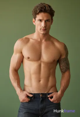 A shirtless man with wavy brown hair stands against a plain grayish-green wall d