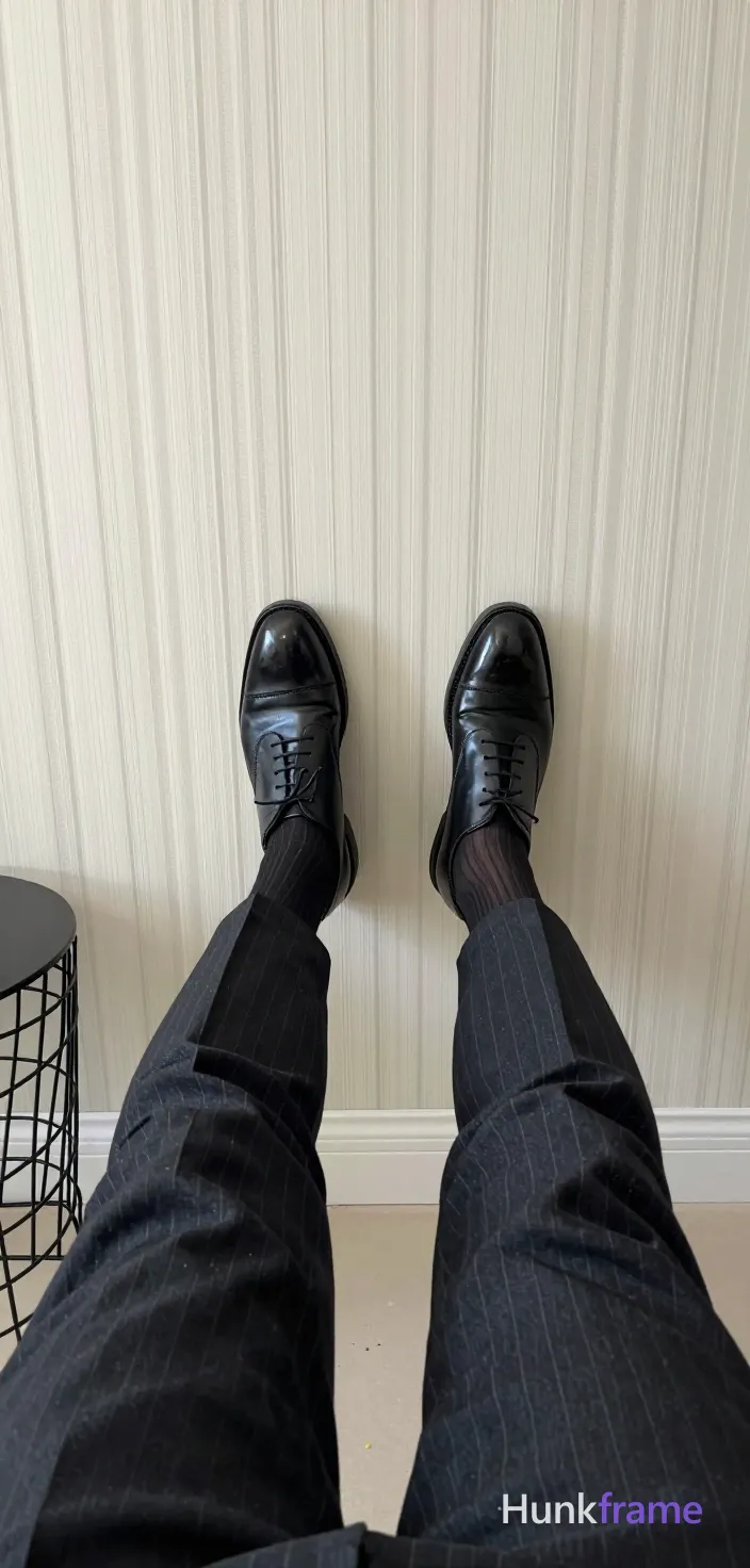 Full-bleed vertical wallpaper composition of an adult man seated with legs extended forward, feet resting on a cream surface. Low-angle camera looking up between thighs to showcase thick leg muscles and dark pinstripe suit trousers from waist to ankles. Black cap-toe oxfords with laces and dark ribbed socks showing subtle vertical stripes on one leg. Clean negative space around wire-frame side table, light wall paneling, soft indoor ambient lighting., a single uninterrupted photograph only, no written words or typography anywhere in frame, no captions, no app UI, no social-media post layout, no screenshot borders