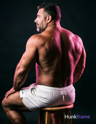 A big hairy stocky adult man with short dark hair and clean-shaven jaw sits shirtless on a wooden st
