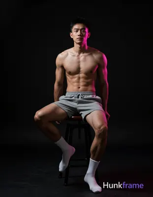 Asian man seated on a dark stool, torso angled away with left leg bent and right extended forward; S