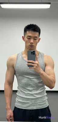 Asian man with short black buzz cut and light stubble stands facing a gym mirror, holding phone for