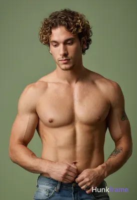 A muscular man with curly hair stands against a plain grayish-green wall during 
