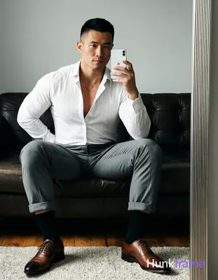 Asian man seated on dark leather sofa, legs spread knees bent taking mirror selfie with white smartp
