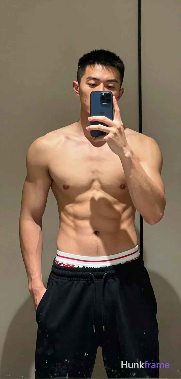Asian man with short black buzz cut and light stubble on jawline, shirtless with defined pectorals, six-pack abs, and visible obliques; Standing facing forward in a mirror reflection, left hand holding blue smartphone up to face for selfie, right hand partially inserted into pocket of black drawstring sweatpants; White briefs with red diagonal stripes visible above waistband over elastic underwear; Indoor bathroom setting with neutral beige walls and vertical black frame lines; Soft frontal fill lighting from phone flash creating even illumination on skin tones against dark pants; Vertical portrait crop centered vertically and horizontally, full torso from upper chest to mid-thighs visible; Premium fashion editorial tone emphasizing sculpted physique; single clean uninterrupted photograph with a cohesive editorial composition and plain continuous surrounding surfaces