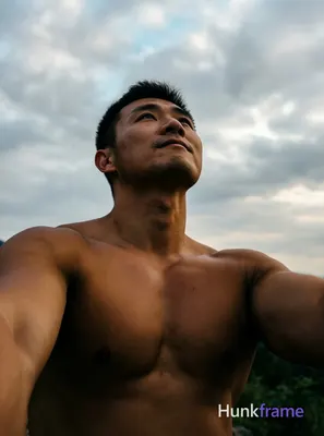 A shirtless man standing with his arms slightly raised, looking upwards towards the sky against an o