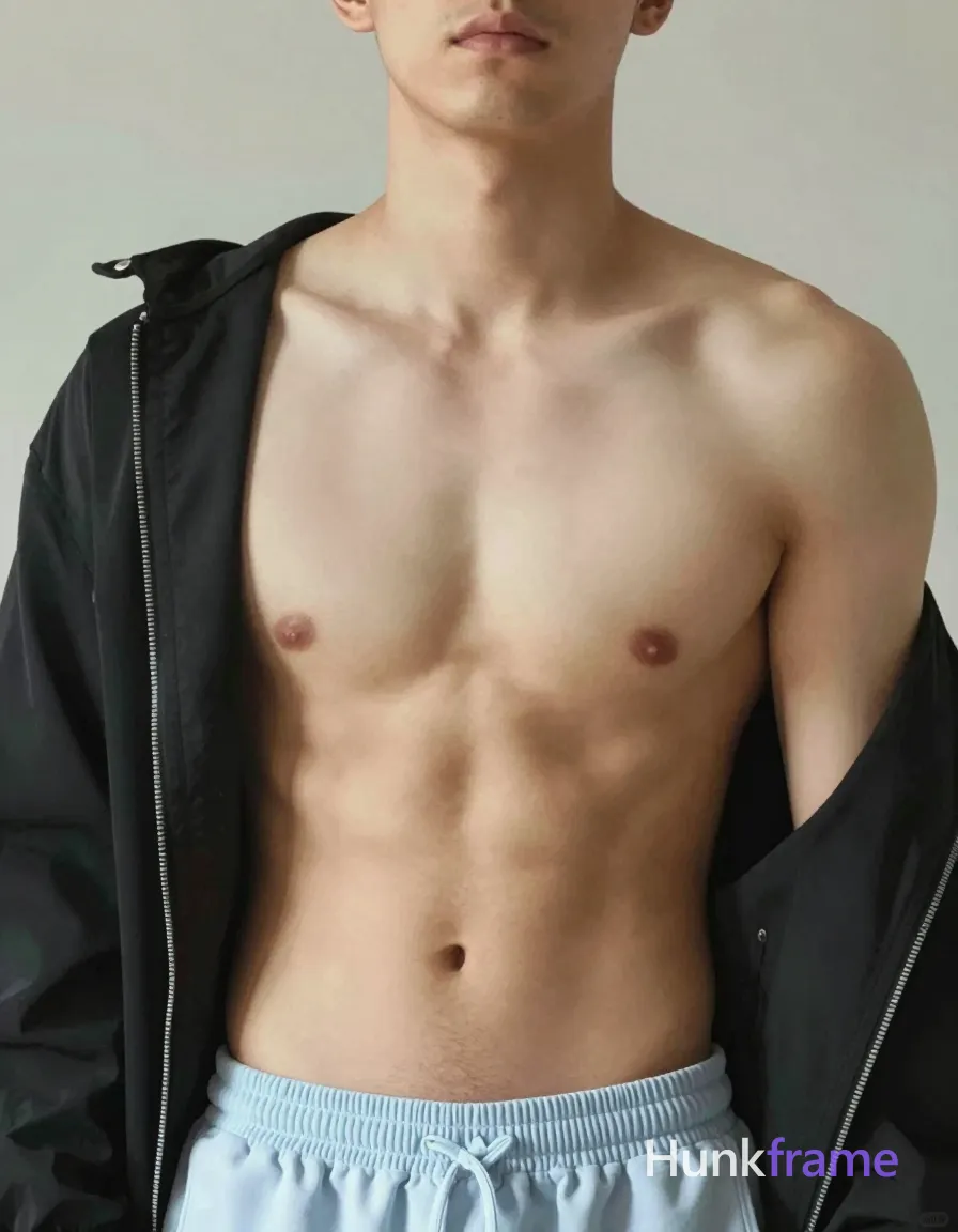 A shirtless man standing with his head slightly tilted forward, wearing a black jacket draped over his shoulders. The focus is on his upper body and torso, highlighting broad shoulders and defined abs. Soft lighting creates a romantic and artistic atmosphere, with the background softly out of focus to draw attention to the subject's muscular form. The image has a neutral palette with subtle film grain, giving it an unretouched photographic quality