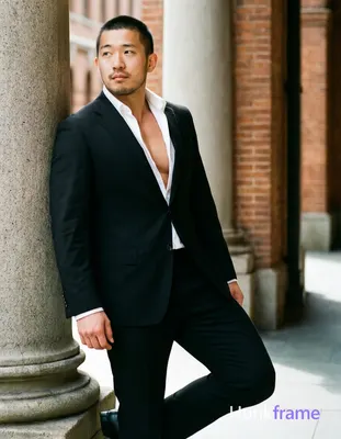 Asian man with short buzz cut and light stubble stands in three-quarter profile leaning against a st