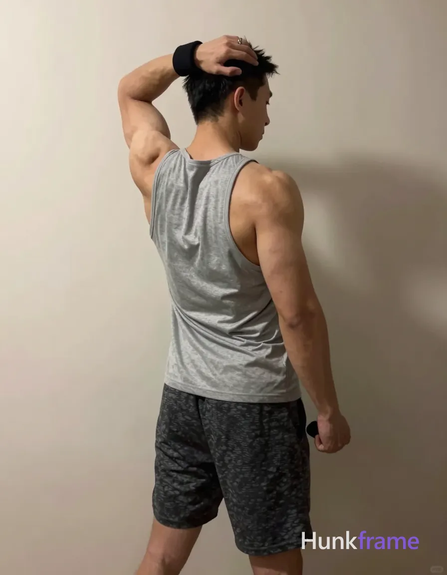 A muscular adult man stands in profile against a plain light-colored wall, grey sleeveless tank top covering torso and back, dark grey textured shorts. His left arm is raised behind his head with elbow bent, gripping a black wristband near the neck; right hand rests by hip holding a black object. Soft directional indoor lighting casts sculpted shadows on defined back muscles and veins on arms. Vertical portrait crop from mid-thighs to upper chest/neck, beige palette, casual fitness-oriented mood., a single uninterrupted photograph only, no written words or typography anywhere in frame, no captions, no app UI, no social-media post layout, no screenshot borders