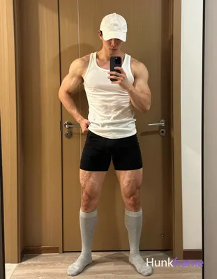 A striking masculine man with short dark hair under a white cap stands in a wide stance against an i