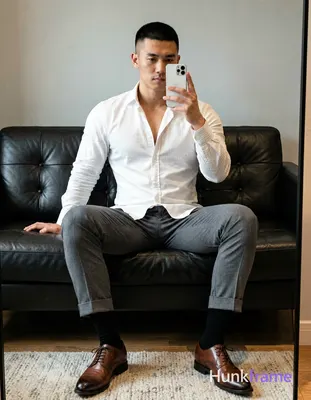 Asian man seated on dark leather sofa, legs spread knees bent taking mirror self